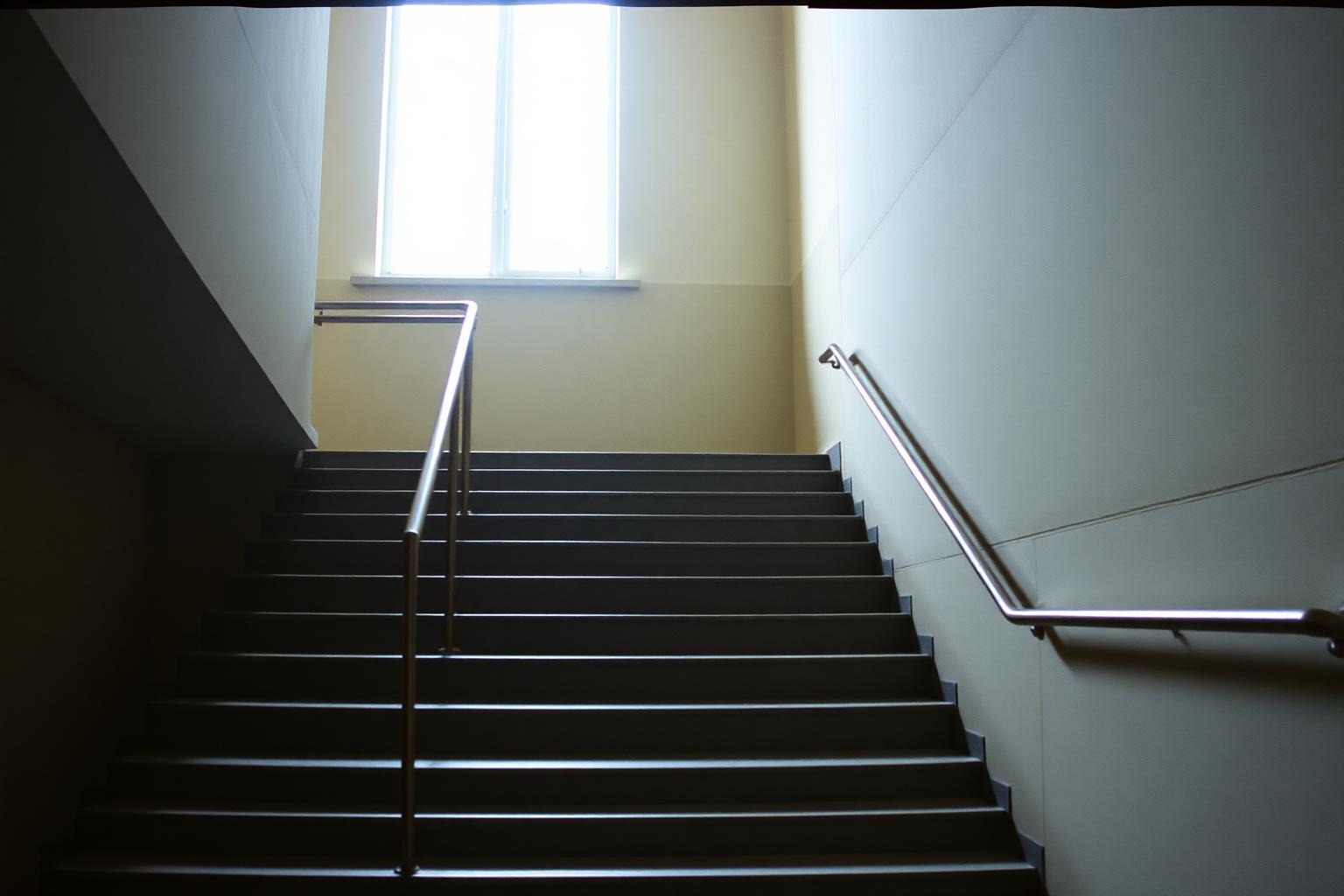 Leere Treppe in einem öffentlichen Gebäude, die zu einem hellen Fenster hinaufführt.
